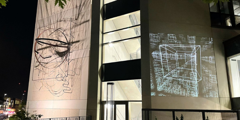 Projection of squares on the side of a building. To the left of the projection is a sculpture of twisted wire also on the building.