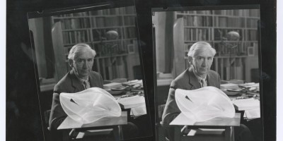 A black-and-white photograph of an older Herbert Read wearing a suit and sat at a desk filled with books and papers in his study at Stonegrave House. In front of him on a small table sits a curved sculpture, made from Perspex and nylon strings. Behind him are a row of bookshelves, a tall wooden sculpture and a wrought iron sculpture.