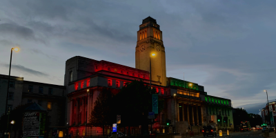 A building with a clocktower and pillars lit with yellow, red and green lights against a dark sky.