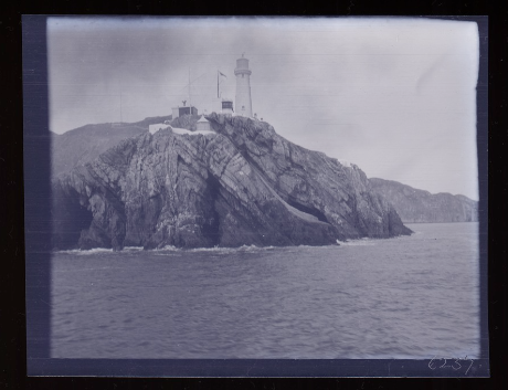 A cellulose nitrate negative photo of a white lighthouse on top of a large rock surrounded by sea. The blue tint gives it a moody feel, creating contrasts between the dark and light shades.
