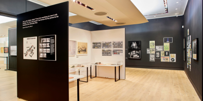 Photograph of the exhibition Another Brick in the Wall. The walls are painted black, on which are black and white photographs and archival images. In the centre of the room is an alcove painted white with glass exhibition cases filled with books.