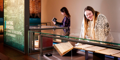 A gallery visitor looks at a museum case containing a historic travel guide book, next to a glowing plinth which says Shifting Borders