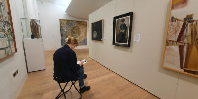 Isabelle, wearing a blue jacket and jeans with her blonde hair pulled up into a hairclip, sat on a black gallery stool sketching on a clipboard in The Stanley & Audrey Burton Gallery