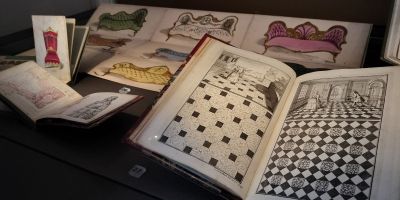 Open books featuring mosaic floor tiles designs and colourful sofa illustrations, in a glass display cabinet.
